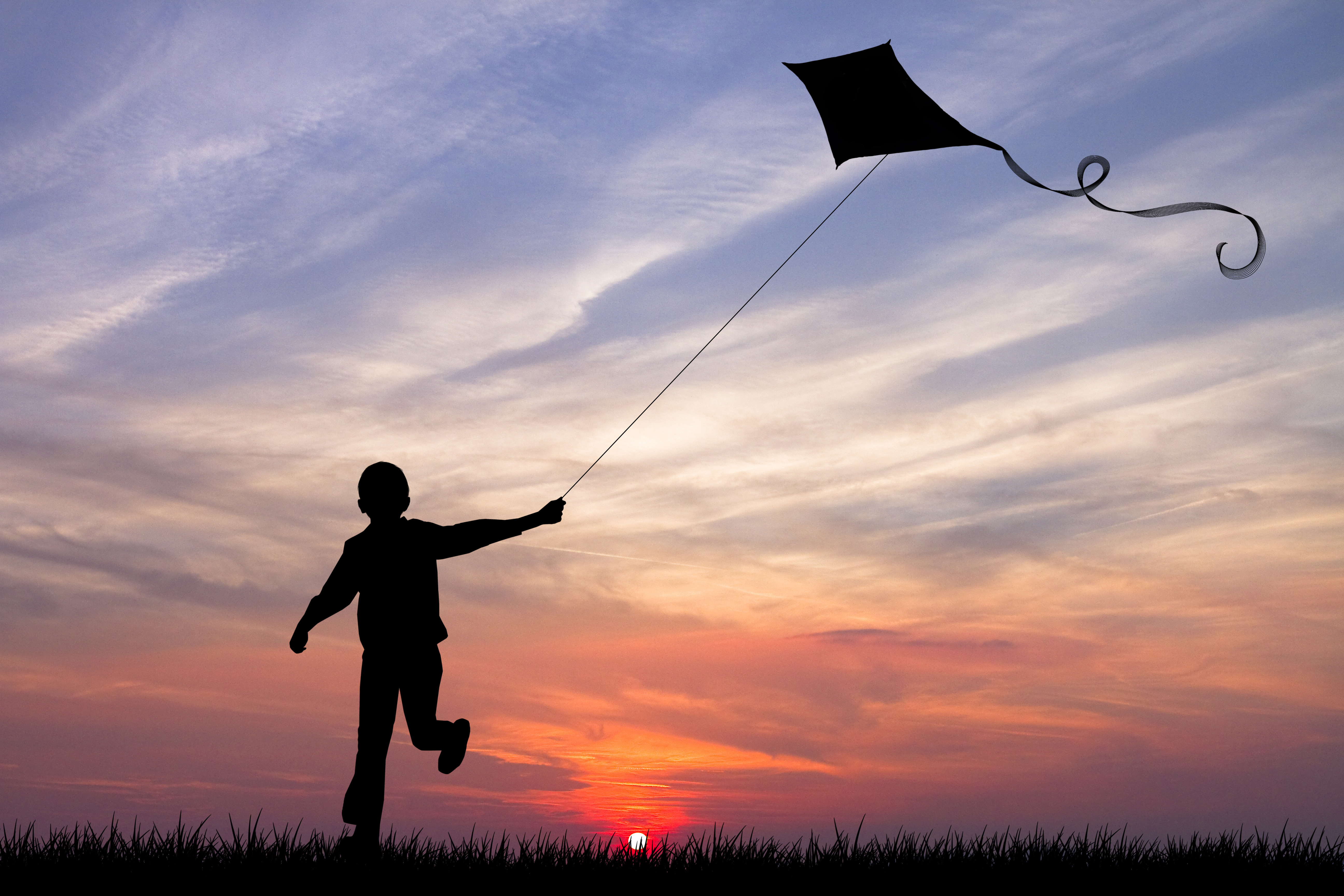 Boy with flying kite running towards the sunset at the horizon